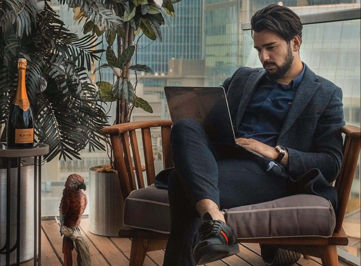 Rich man sitting on a chair in his balcony, with his laptop on his lap.