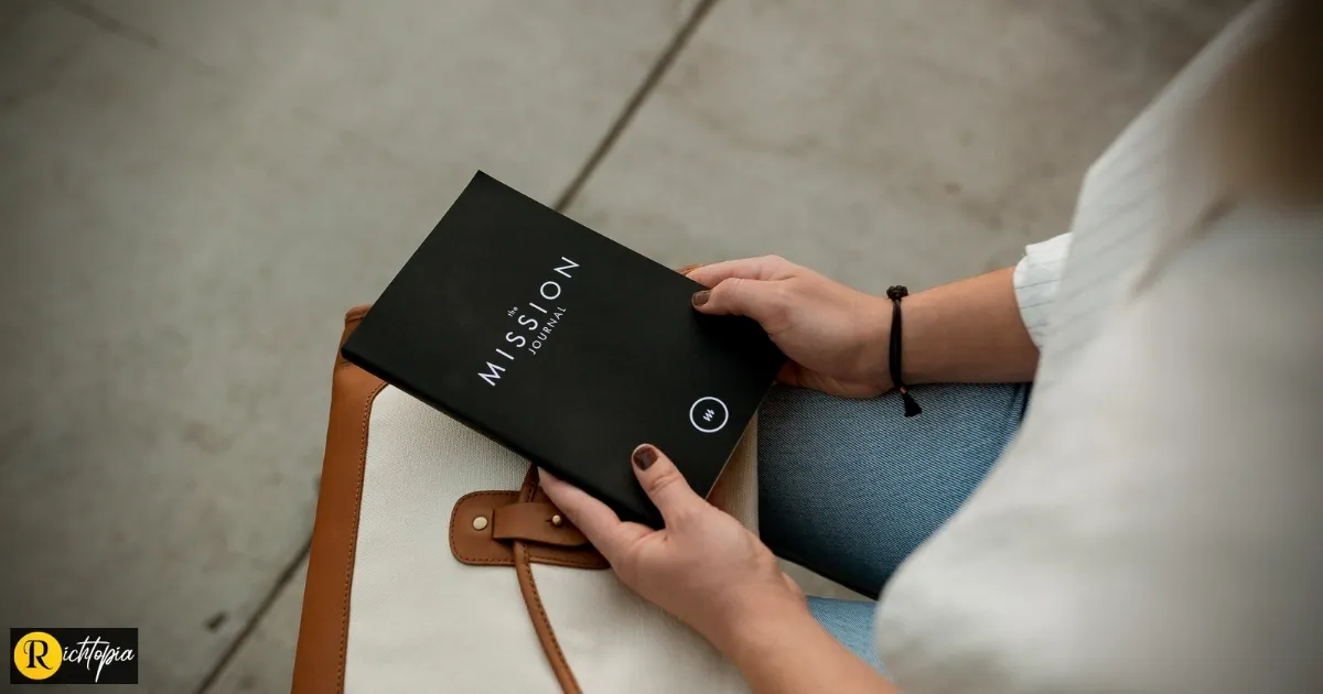 Photo of a woman sitting down with a black coloured diary entitled 'mission' in her hand.