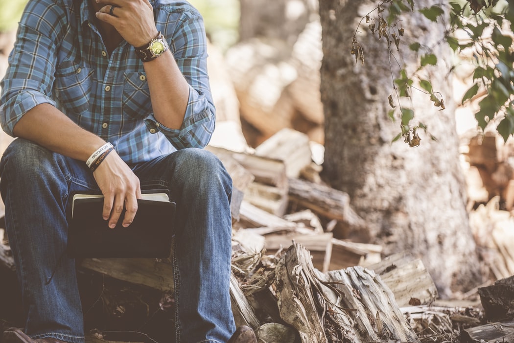 Man sitting in the woods wearing blue plaid dress shirt and blue jeans with a notepad in his hand.