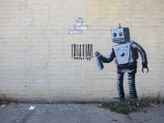Graffiti art of emotionally intelligent robot spray-paining on a brick wall in the street.