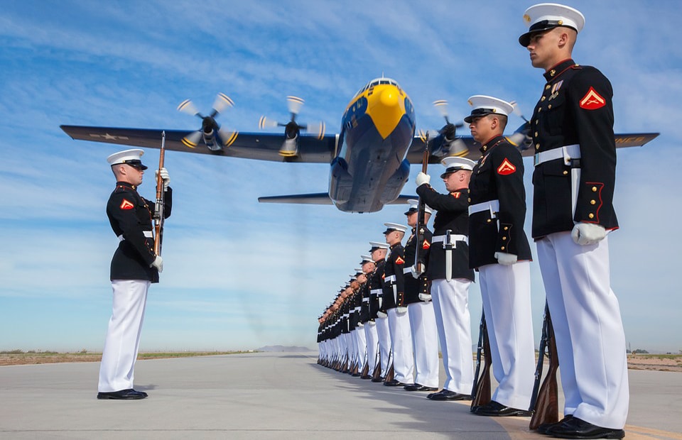 Photo of Marines, who serve the USA, conducting a silent drill and a military plane flying close-by in the background.