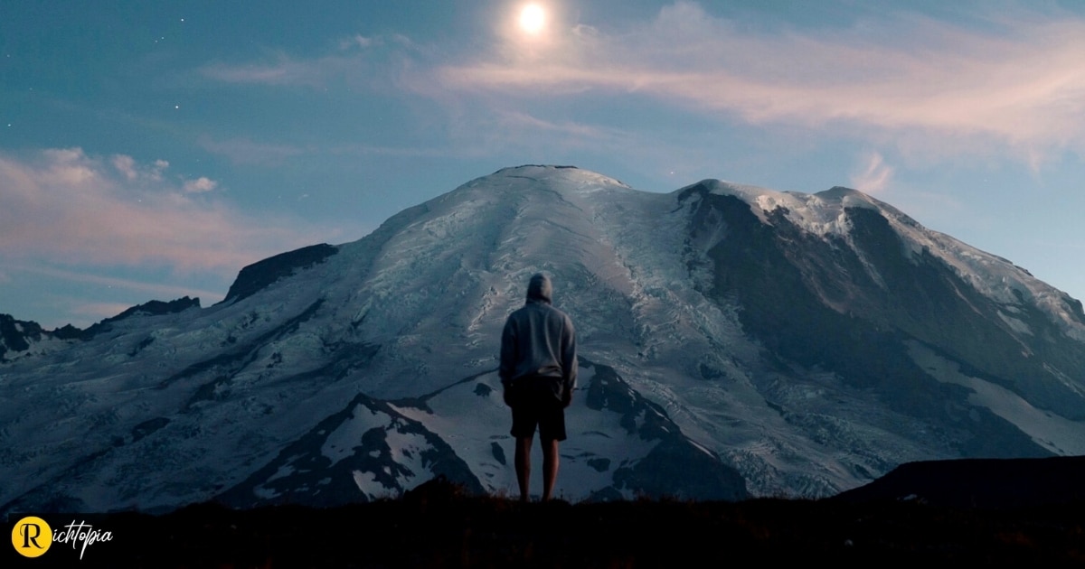 Photo of a man wearing a hoodie watching a picturesque sunset over an icy mountain in front of him.