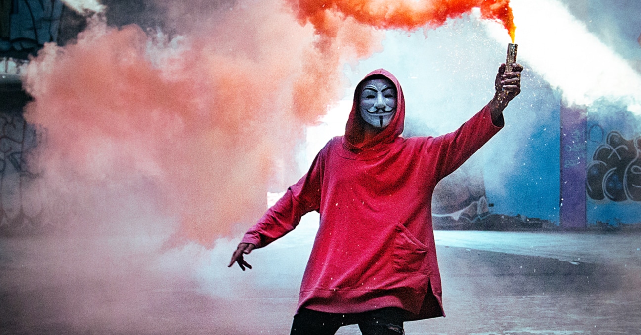 Photo of person in guy-fawkes mask, with a red hoodie, holding a red smoke torch. Article about IoT security threats and risks.