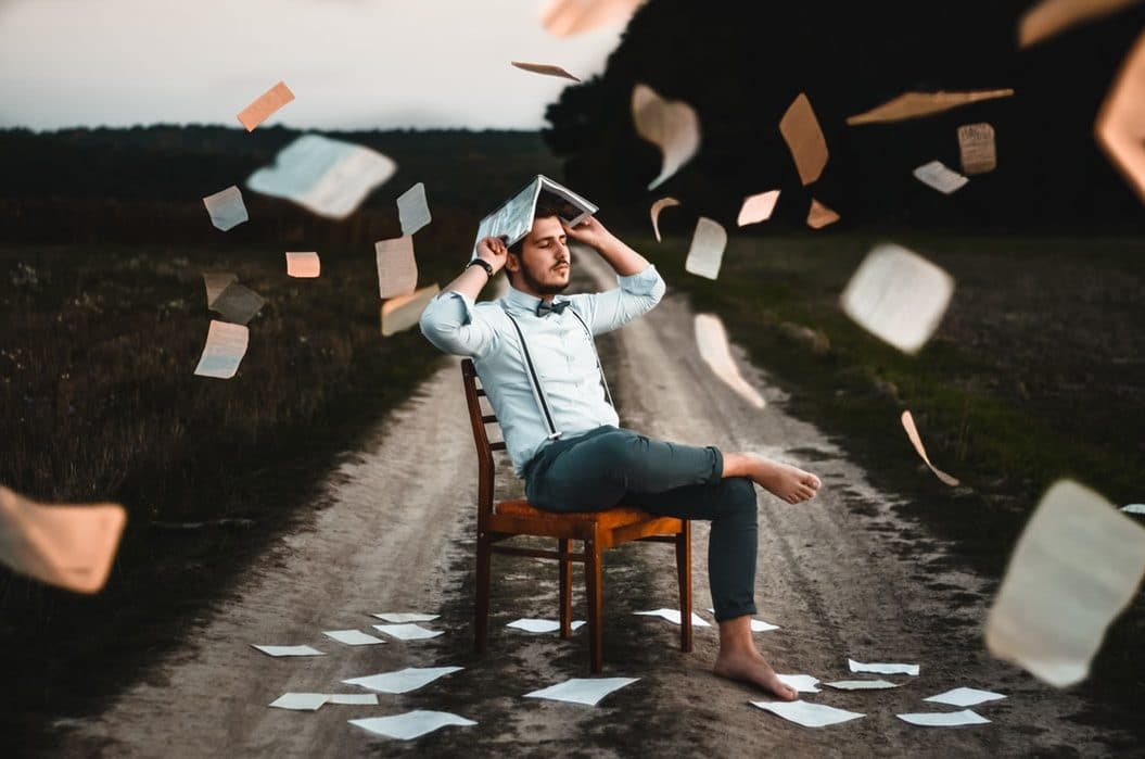 Man sitting on a chair in the middle of a dirt road with a book on his head and pieces of paper flying around.