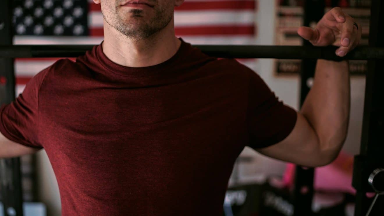 Strong man holding a barbell on his shoulders, in a room with a US flag in the background.