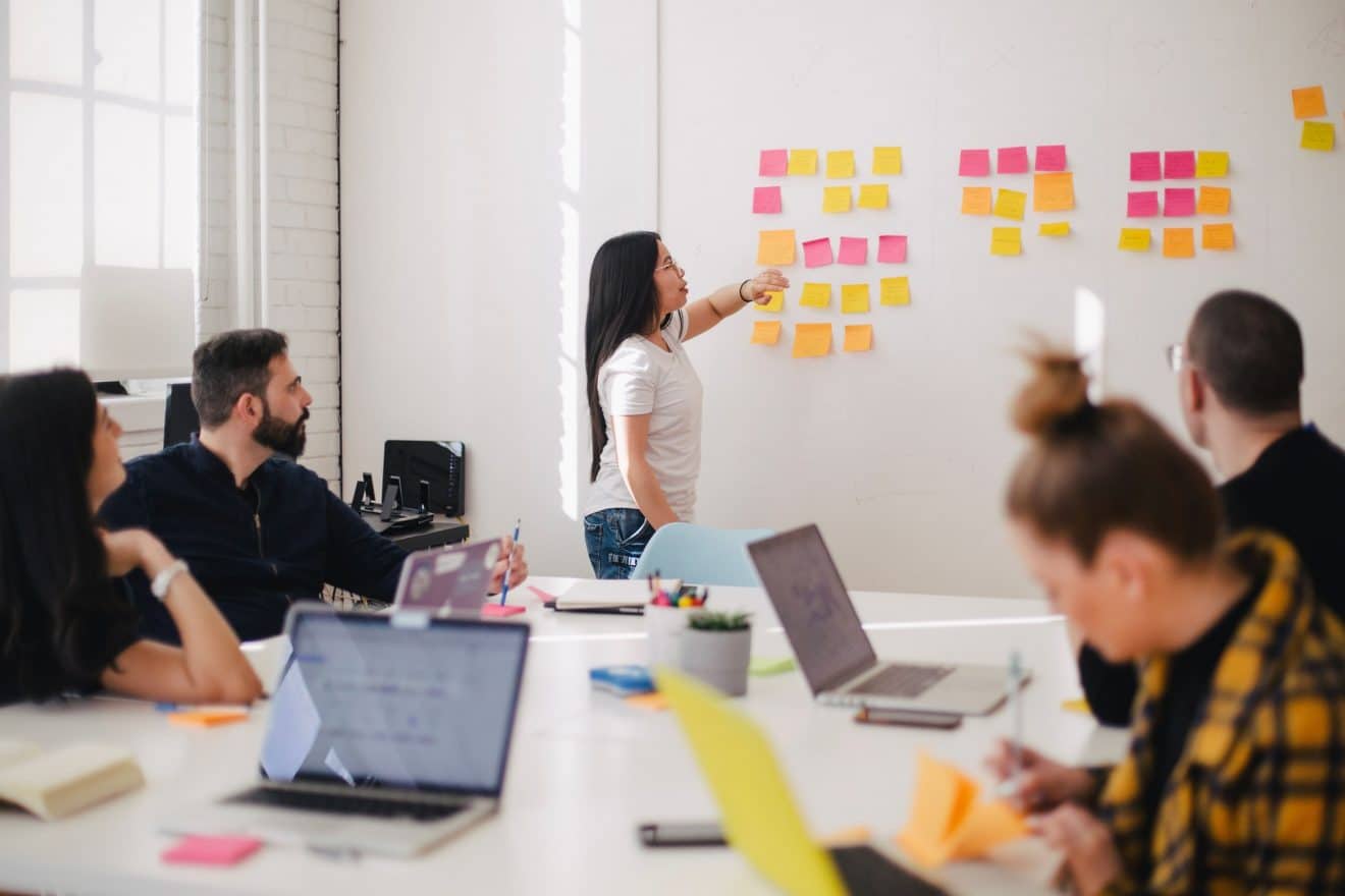 Woman placing sticky notes on a whiteboard during a business meeting whilst people sitting on a table with their laptops take notes.