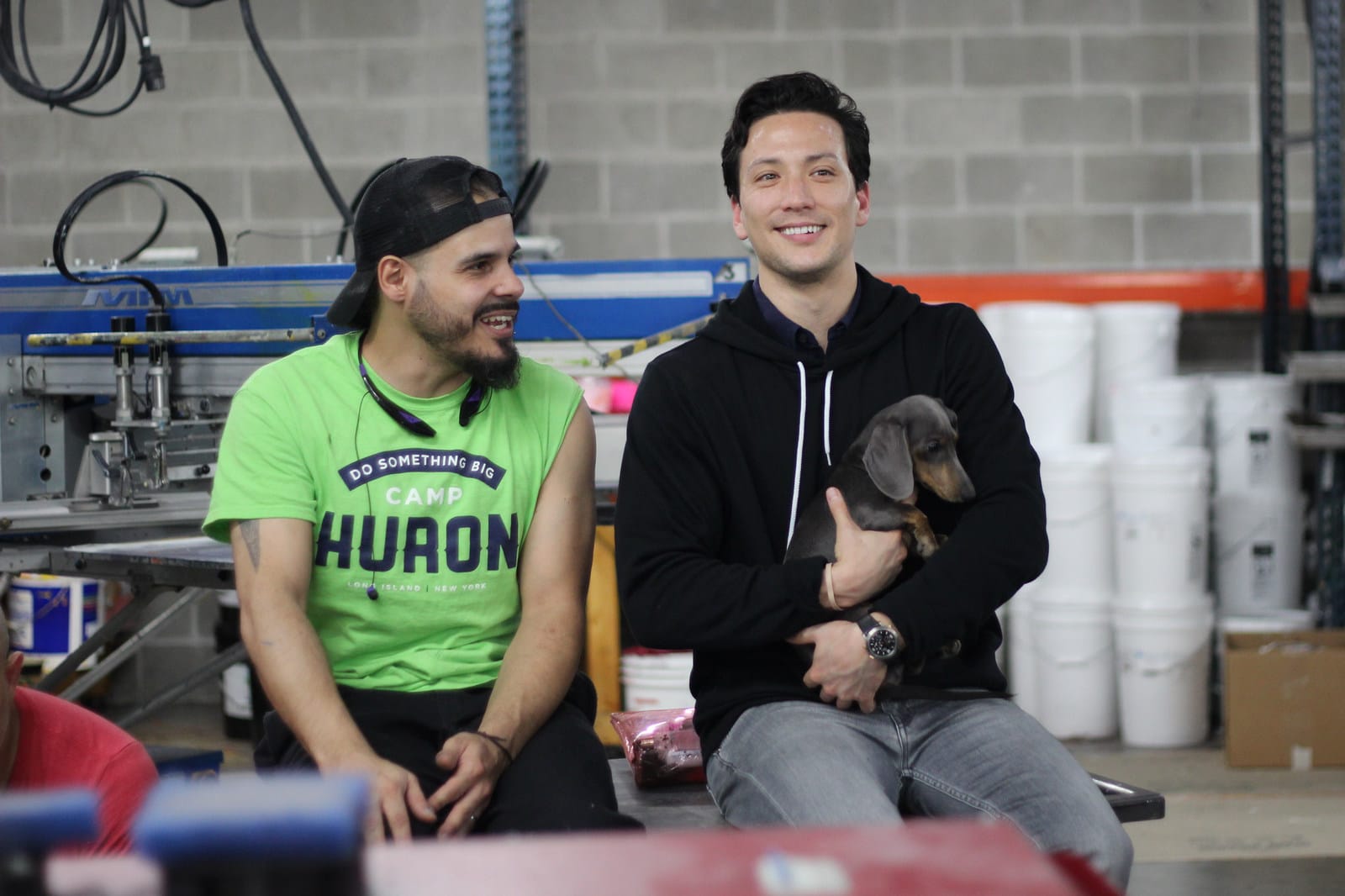 Michael Nemeroff sitting on a work table beside his employee at RushOrderTees factory holding his dog on his lap