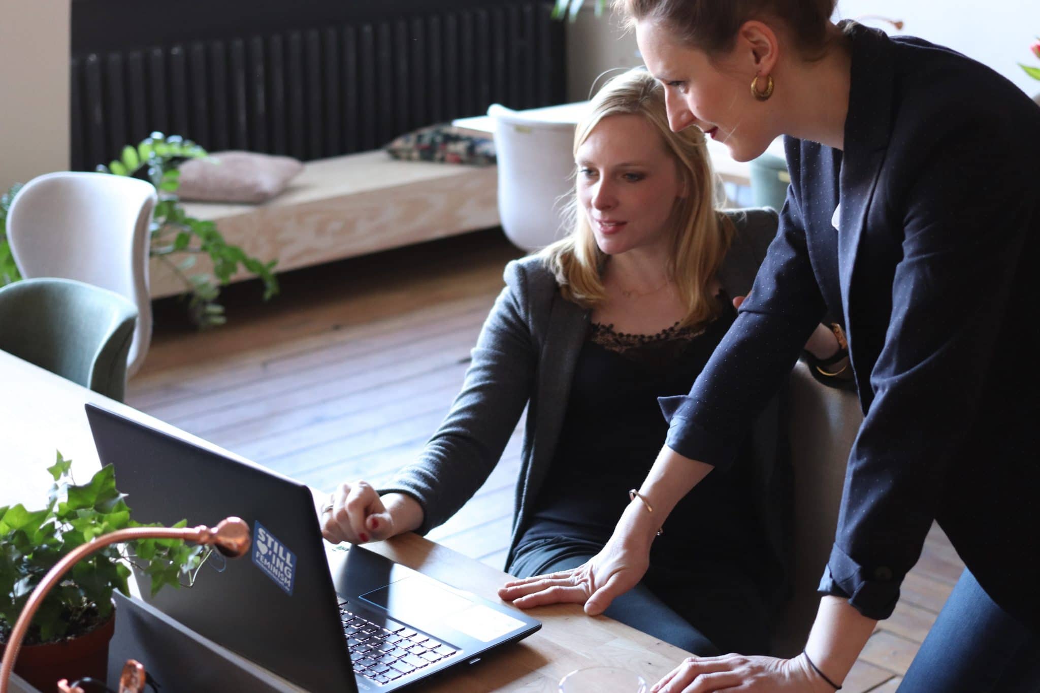 Photo of two women, one sitting on a desk, whilst the other stands behind her. They are both looking into a laptop with excitement in their faces.