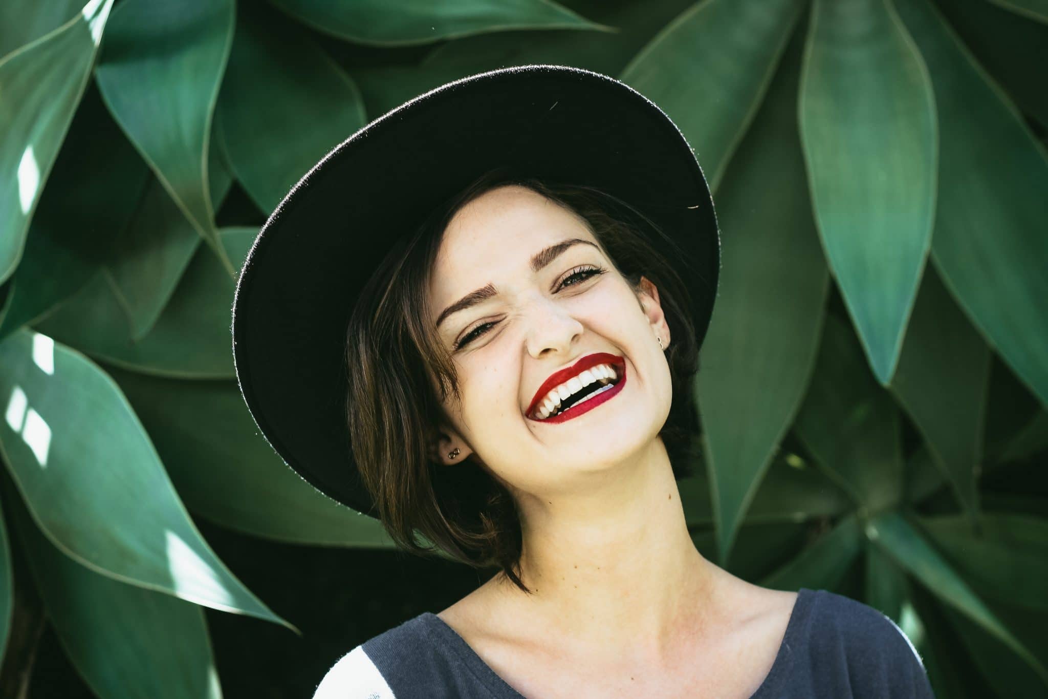 Photo of a woman wearing a stylish hat and smiling whilst standing in front a green plant.