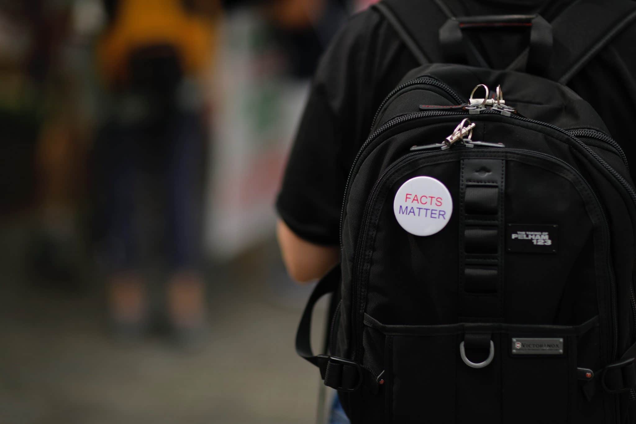 Photo of a man wearing a backpack, taken from the back, with a pinned logo on the bag stating, 'facts matter'.