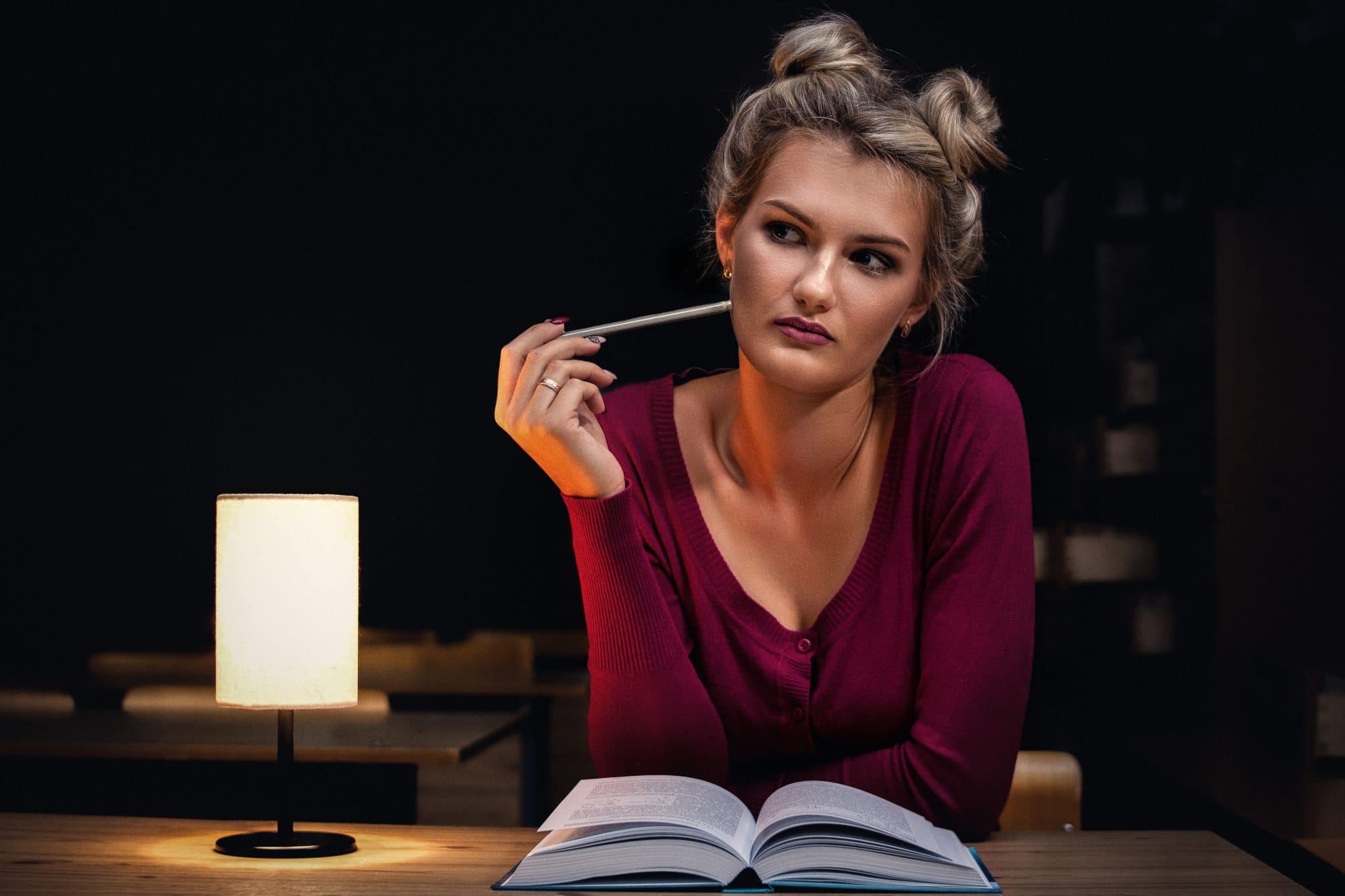 Woman sitting on a library desk beside opened book holding pencil to her cheek whilst thinking, with a beautiful lamp in the sideground.