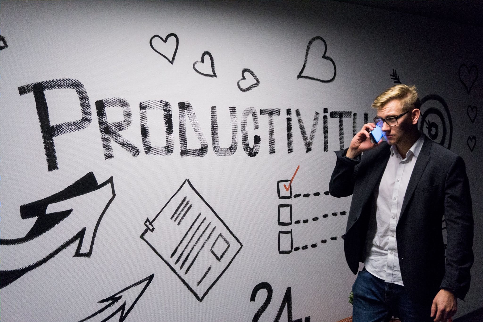 Photo of a blonde man in suit wearing glasses taken as he talks on his smartphone. In the background there is an white plasterboard with the word 'productivity' painted onto it, along with shapes of hearts and other symbols.