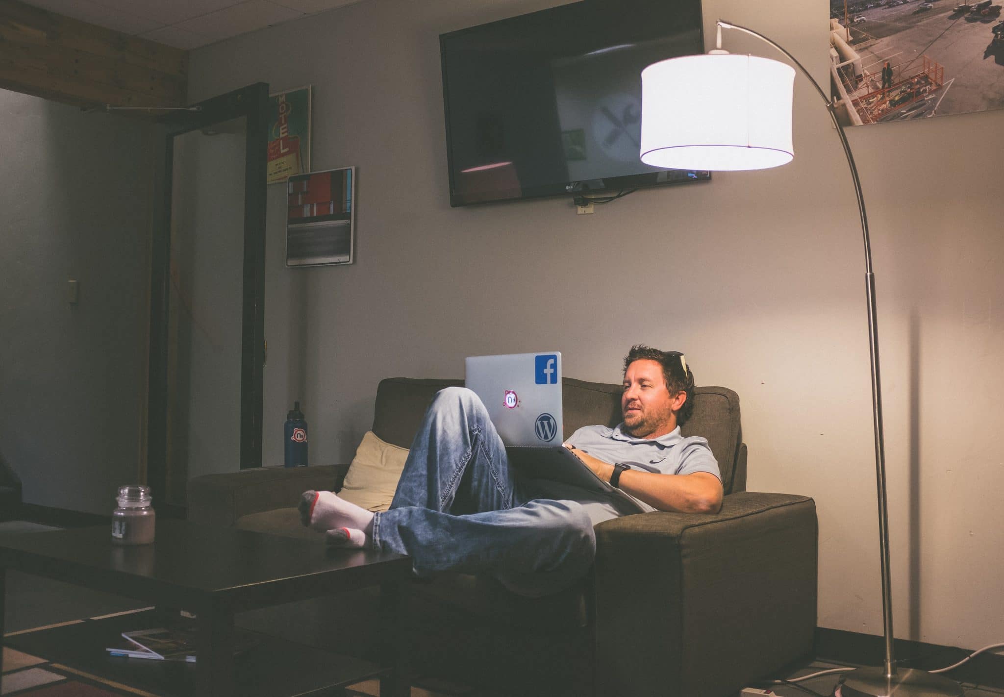 Photo of a man on his laptop with stickers of facebook and wordpress whilst slouching on a couch in his living room