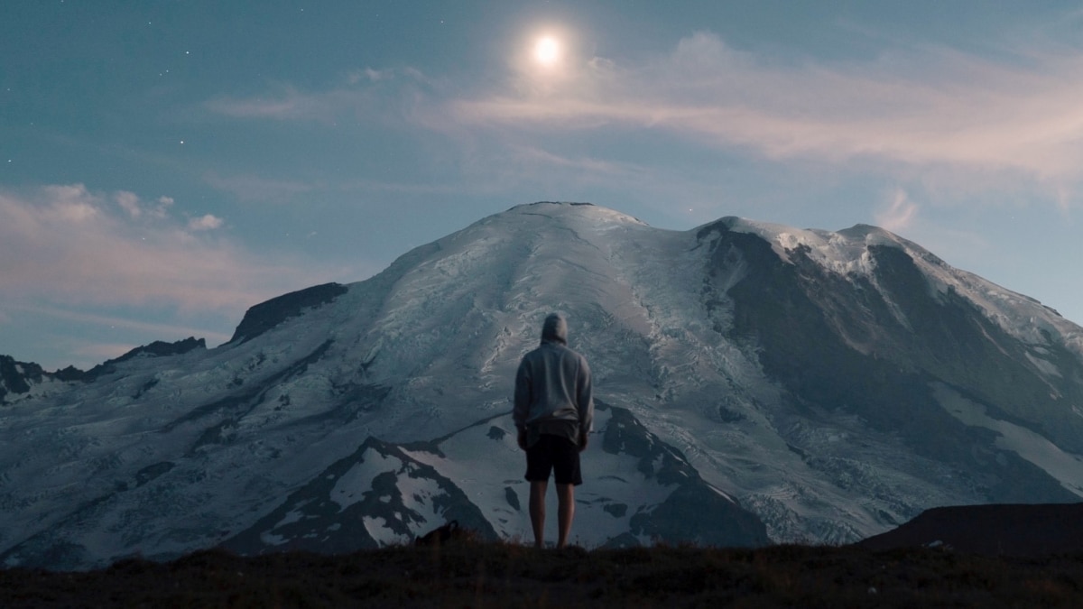 Photo of a man looking into the sunset on top of a big mountain in front of him.